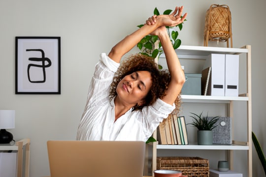 A woman stretching while working using laptop at a home office.