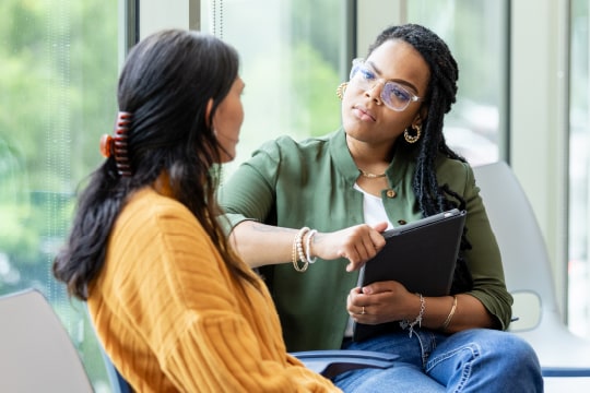 A woman seated and listening attentively to another woman, while holding a tablet.