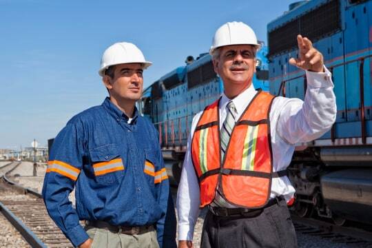 Two professionals in hardhats standing in a railroad construction site, one pointing as both look out into the distance beyond the camera.
